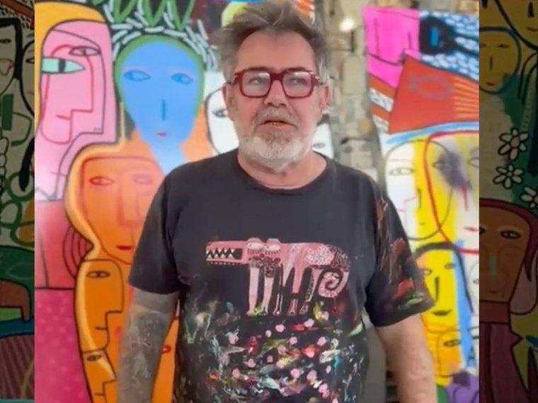 El artista Milo Lockett frente a un mural colorido lleno de rostros.